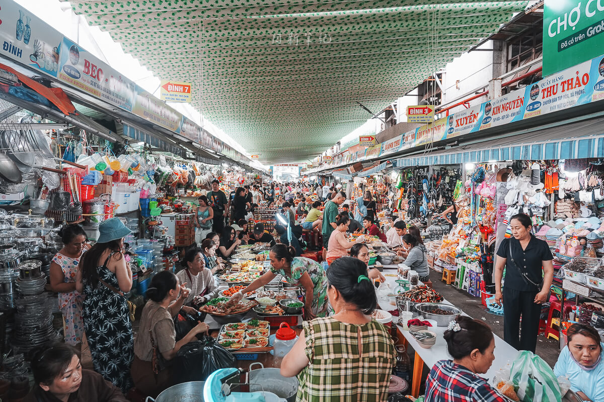 峴港共市場雜貨攤位，擺滿紅色罐子、擺飾與彩繪斗笠，旁邊一位客人在看商品