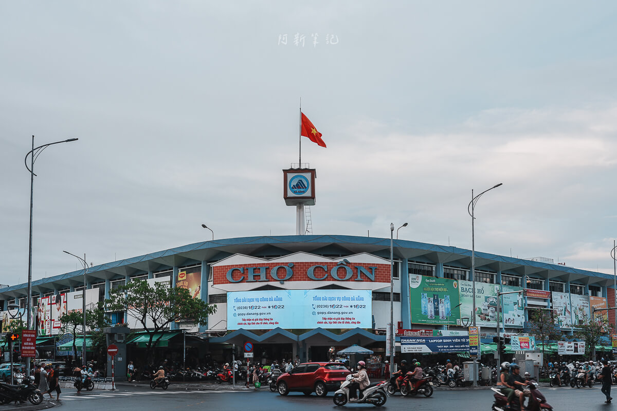從街道拍攝峴港共市場建築全景，屋頂鐘塔上的越南國旗與門前車流